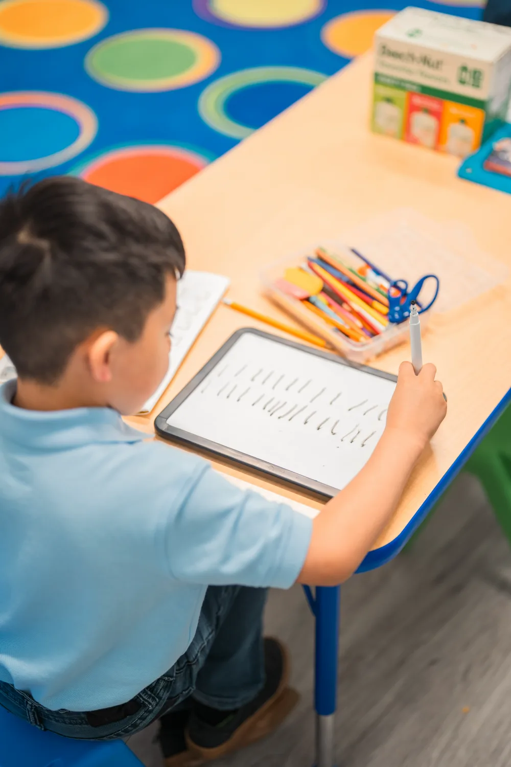 A student practising handwriting strokes on a small whiteboard at a classroom desk.