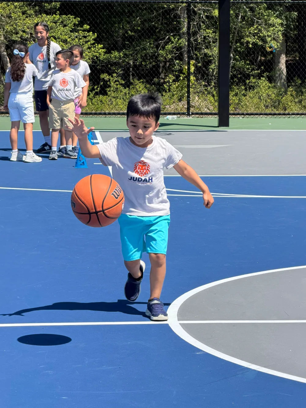 A student dribbling a basketball on an outdoor court, wearing the white activity shirt with the lion logo.
